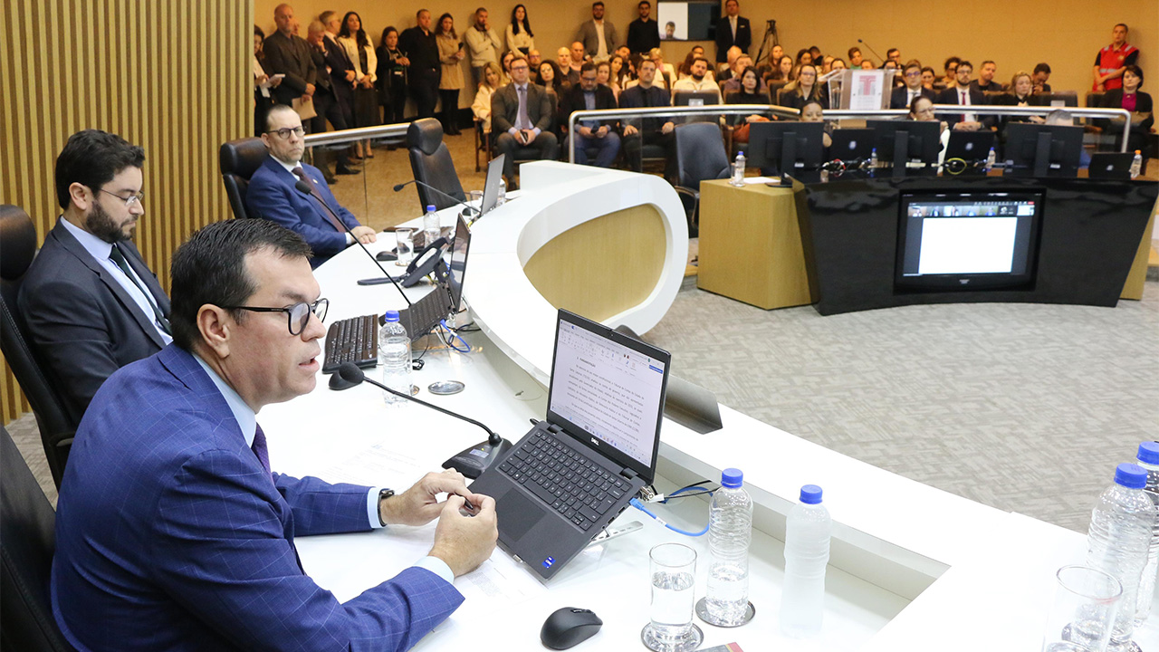 Banner horizontal com foto de perfil do conselheiro Adircélio de Moraes durante a sessão plenária de apreciação das contas. Ele é um homem branco, de cabelos castanhos. Está sentado diante da bancada de autoridades, veste terno azul, usa óculos de grau e fala ao microfone. Ao seu lado, aparecem outras autoridades. Ao fundo, em poltronas, a plateia que assiste à sessão.