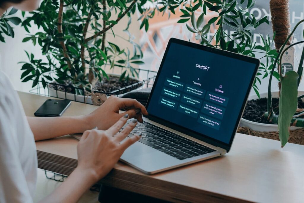 A imagem mostra uma pessoa usando um laptop em uma mesa de madeira, com a tela exibindo a interface do ChatGPT. Ao fundo, há plantas decorativas, criando um ambiente natural e agradável. Um celular está sobre a mesa ao lado do laptop. As mãos da pessoa estão no teclado, indicando que está interagindo com o computador.