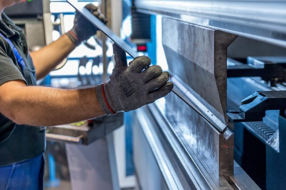Fotografia colorida mostra um trabalhador operando uma máquina industrial de dobra de metal. A pessoa veste camiseta preta, avental azul e luvas de proteção cinza, segurando uma chapa metálica com ambas as mãos. A cena ocorre em um ambiente industrial iluminado, com detalhes de equipamentos ao fundo, evidenciando o cuidado e a precisão no manuseio das peças.