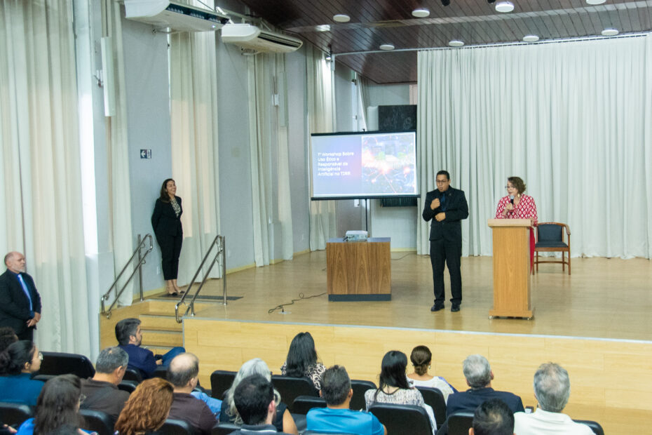 A imagem colorida mostra um auditório durante um workshop. Em primeiro plano, a parte de trás da plateia é visível, com várias pessoas sentadas em cadeiras estofadas escuras, voltadas para o palco. No palco, que é de madeira clara, uma mulher e um homem estão de pé. O homem, à esquerda, o interprete de Libras, enquanto a mulher, à direita, está em frente a um púlpito. Há dois púlpitos de madeira no palco, um ao lado do outro, e um banco de madeira mais baixo entre eles. Ao fundo do palco, cortinas claras cobrem a parede.