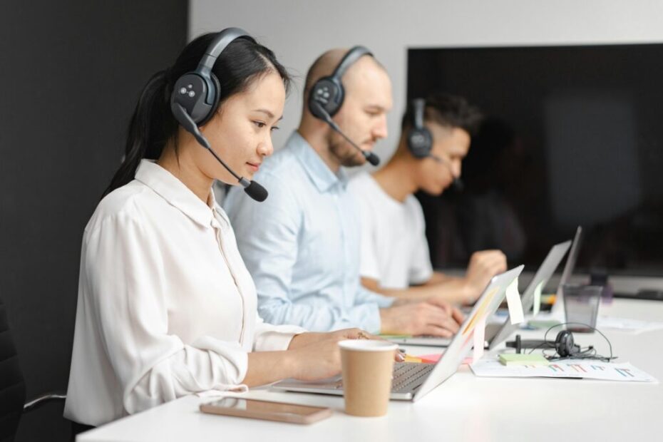 A imagem mostra três pessoas sentadas em uma fileira em frente a computadores, usando fones de ouvido com microfone. Elas parecem estar trabalhando em um ambiente de escritório, possivelmente em um call center ou em um ambiente de atendimento ao cliente. A pessoa à frente está mais focada, enquanto as outras duas estão levemente desfocadas, criando um efeito de profundidade de campo. As pessoas estão usando camisas sociais e o ambiente parece profissional e organizado. A iluminação é suave e neutra. Há papéis e canetas na mesa, sugerindo trabalho administrativo ou de atendimento. Um grande monitor está no fundo, mas sua tela está escura e não mostra detalhes. A imagem transmite uma sensação de trabalho em equipe e foco em tarefas.