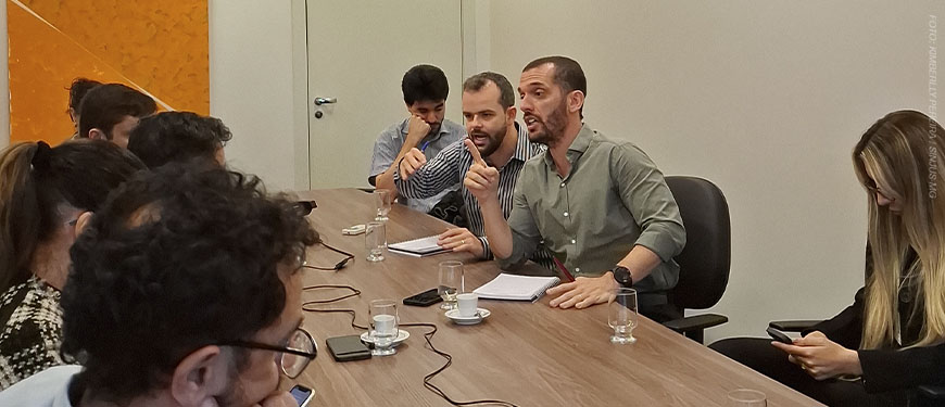 Reunião com cerca de dez pessoas sentadas ao redor de uma mesa, em ambiente interno e iluminado. No centro da imagem, há dois dirigentes do SINJUS-MG: o coordenador-geral, Alexandre Pires e o diretor de Assuntos Jurídicos, Felipe Rodrigues. Ambos falam com expressividade e gesticulam enquanto seguram papéis. Os demais participantes observam atentamente ou fazem anotações.