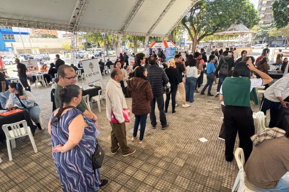 A imagem mostra um evento ao ar livre, possivelmente uma feira de empregos ou um evento comunitário, sob uma grande tenda branca. Há uma multidão de pessoas em pé e sentadas, interagindo e se movendo pelo espaço. Várias mesas com materiais informativos e banners estão dispostas, com algumas pessoas em pé atrás delas, possivelmente representando organizações ou oferecendo informações. O ambiente parece ser urbano, com edifícios e árvores visíveis ao fundo. A iluminação sugere que é dia, com luz solar entrando pela lateral da tenda.