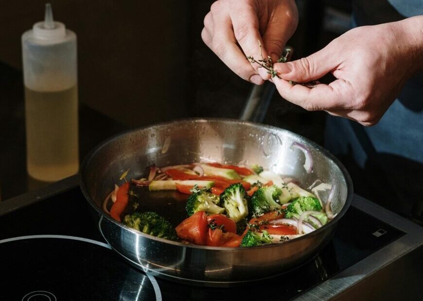 Na imagem aparece uma mão0 segurando uma erva, para colocar nos legumes que estão na frigideira, onde aparecem, brócolis, pimentão vermelho, cebola, tomate e cenoura.