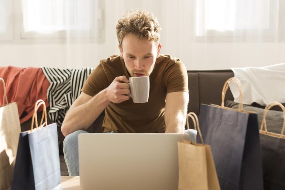 Rapaz em frente ao computador, com sacos de papel à sua volta (a representar compras online). Está a segurar uma chávena