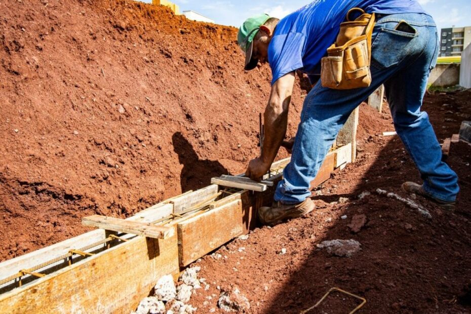 A imagem mostra um trabalhador da construção civil em um canteiro de obras, concentrado em seu trabalho. Ele está agachado, ajustando ou fixando uma estrutura de madeira que serve como forma para o concreto da fundação. O trabalhador veste uma camiseta azul, calça jeans, boné verde e botas de trabalho marrons, além de um cinto de ferramentas de couro na cintura. Atrás dele, há uma grande pilha de terra vermelha escavada. Ao fundo, é possível ver um prédio moderno de vários andares sob um céu azul claro, indicando que a cena ocorre durante o dia. A iluminação é natural e forte, destacando as cores e os detalhes da cena. A imagem transmite o início da construção, focando na preparação da fundação para a edificação.