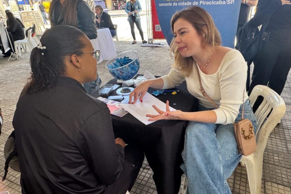 A imagem mostra um cenário ao ar livre, provavelmente em um evento de recrutamento ou feira de empregos. Duas mulheres estão sentadas frente a frente em uma mesa coberta por uma toalha preta. A mulher à direita, loira, veste uma blusa de manga comprida clara e calça jeans azul clara, e está falando, gesticulando com as mãos. A mulher à esquerda, de cabelos curtos e escuros, usa uma blusa preta e óculos, e está ouvindo atentamente. Sobre a mesa há uma tigela transparente com balas embrulhadas em papel azul e alguns papéis. Ao fundo, é possível ver outras pessoas em pé e um banner azul com texto em português que menciona vagas de emprego na cidade de Piracicaba/SP. A cena transmite uma atmosfera profissional e informal, com luz natural do dia.